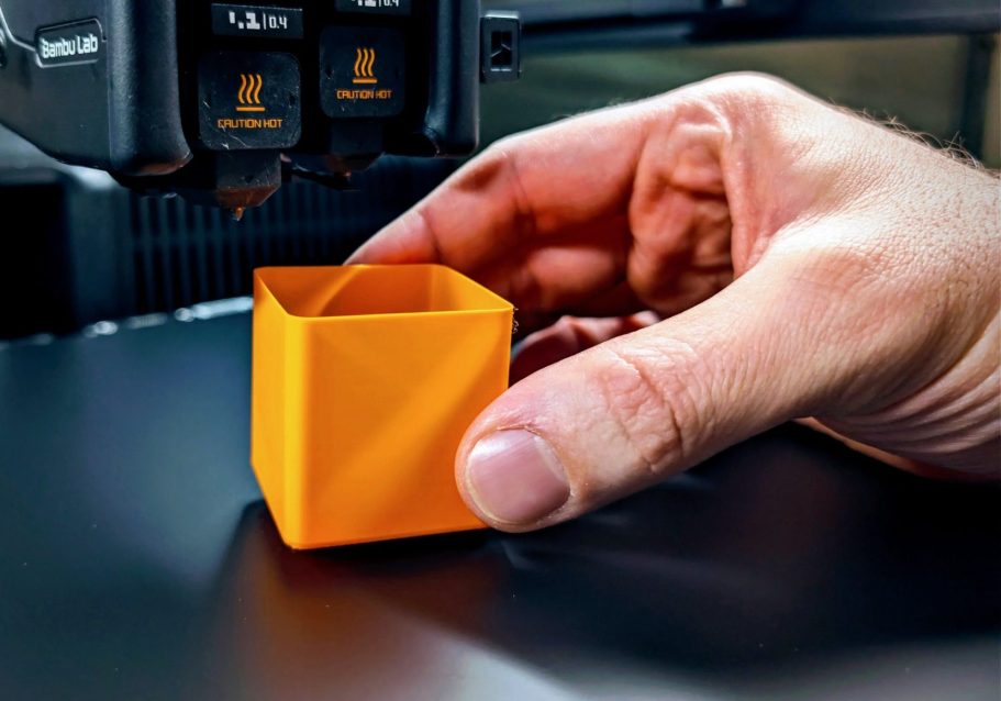Warum Schäfer3D Eine Hand hält einen orangefarbenen, quaderförmigen Behälter neben einem 3D-Drucker.