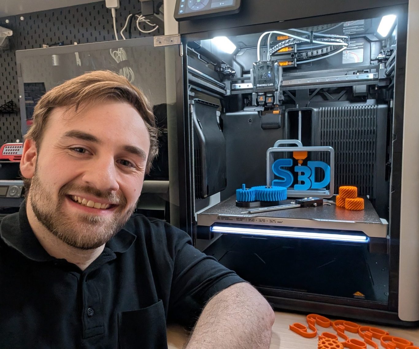 Inhaber | Patrick Schäfer | Schäfer3D Patrick Schäfer lächelt neben einem 3D-Drucker mit bunten Druckobjekten.